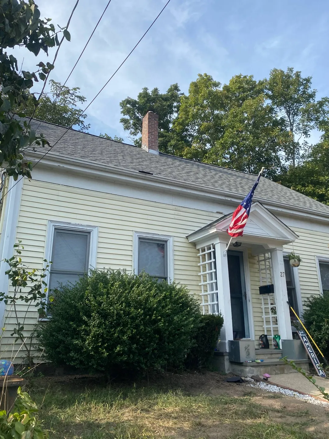 A cream-colored house with a small porch and an American flag hanging by the door under a blue sky.